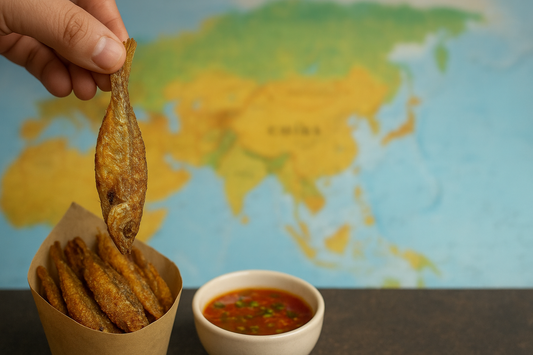 A close-up of crispy fried dry fish served in a paper cone with a small bowl of dipping sauce in the foreground, set against a blurred map of Asia in the background, symbolizing Asian street food culture