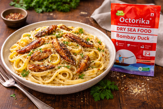 MasterChef-style wooden table setup with a bowl of creamy pasta topped with crispy dry fish chunks and fresh herbs, placed alongside an Octorika Bombay Duck dry seafood packet
