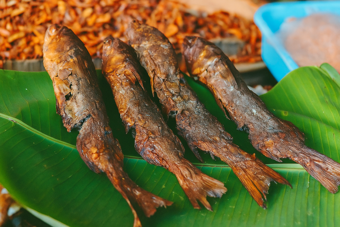 Four whole dry fish placed on a fresh green banana leaf, showcasing their crispy, sun-dried texture with charred brown skin. The background includes dried seafood and market elements, creating a traditional South Indian coastal market vibe.