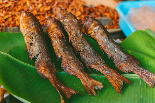Four whole dry fish placed on a fresh green banana leaf, showcasing their crispy, sun-dried texture with charred brown skin. The background includes dried seafood and market elements, creating a traditional South Indian coastal market vibe.