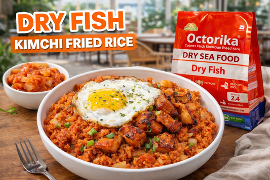 A bright open-restaurant style food photograph showing a bowl of Dry Fish Kimchi Fried Rice topped with crispy dry fish chunks and a sunny-side-up egg, served on a wooden table with a side of kimchi and an Octorika Dry Fish packet in natural daylight