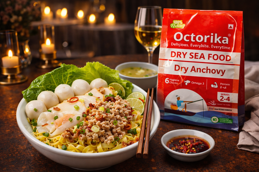 Dry Fish Noodle Bowl topped with crispy dry anchovy, minced fish, silky fish fillet, fish balls, fresh greens, and lime wedges, served on a candle-lit table beside an Octorika Dry Anchovy packet with dipping sauce and chopsticks