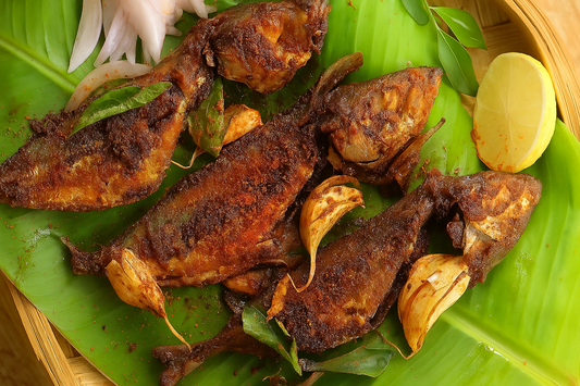 A plate of crispy fried dry fish served on a fresh banana leaf, garnished with roasted garlic, curry leaves, sliced onions, and a lemon wedge, presented in a traditional bamboo tray.
