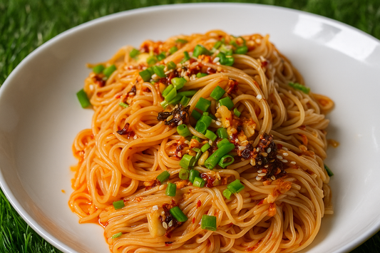A plate of Korean-style noodles served on a white dish, topped with spicy garlic chili oil, finely chopped green onions, sesame seeds, and crispy fried bits. The background features a natural green grass texture